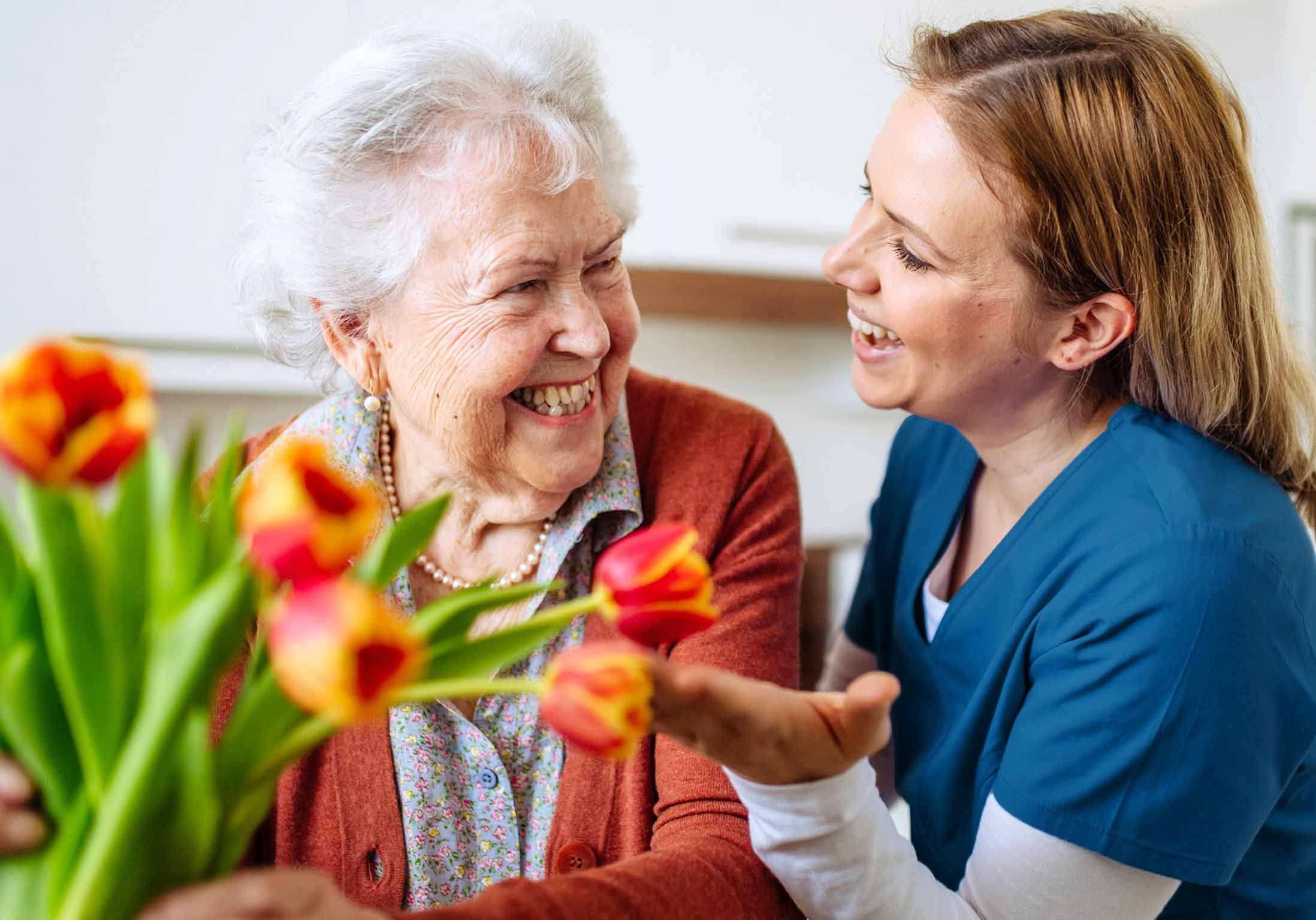 une femme âgée tient des fleurs avec son aide à domicile qui lui sourit